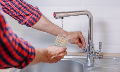 man-pouring-glass-of-water-from-tap-with-clean-filter-in-kitchen-up-picture-id1050746464-min